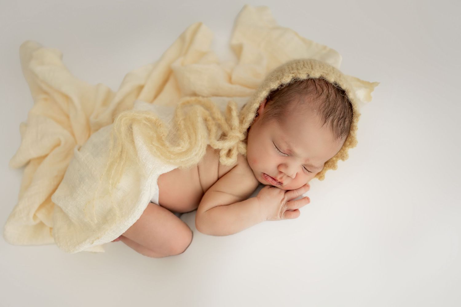 Sleeping baby swaddled in white wrap with tan bow headband on cream shag rug.
