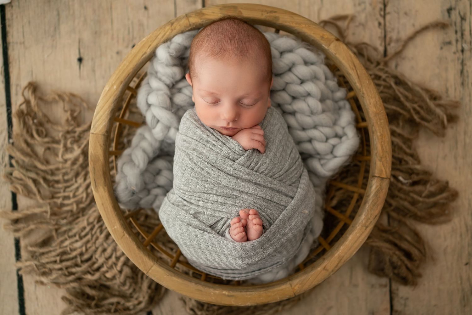 Newborn baby swaddled in soft grey knit wrap sleeping peacefully in wooden bowl on rustic burlap background.