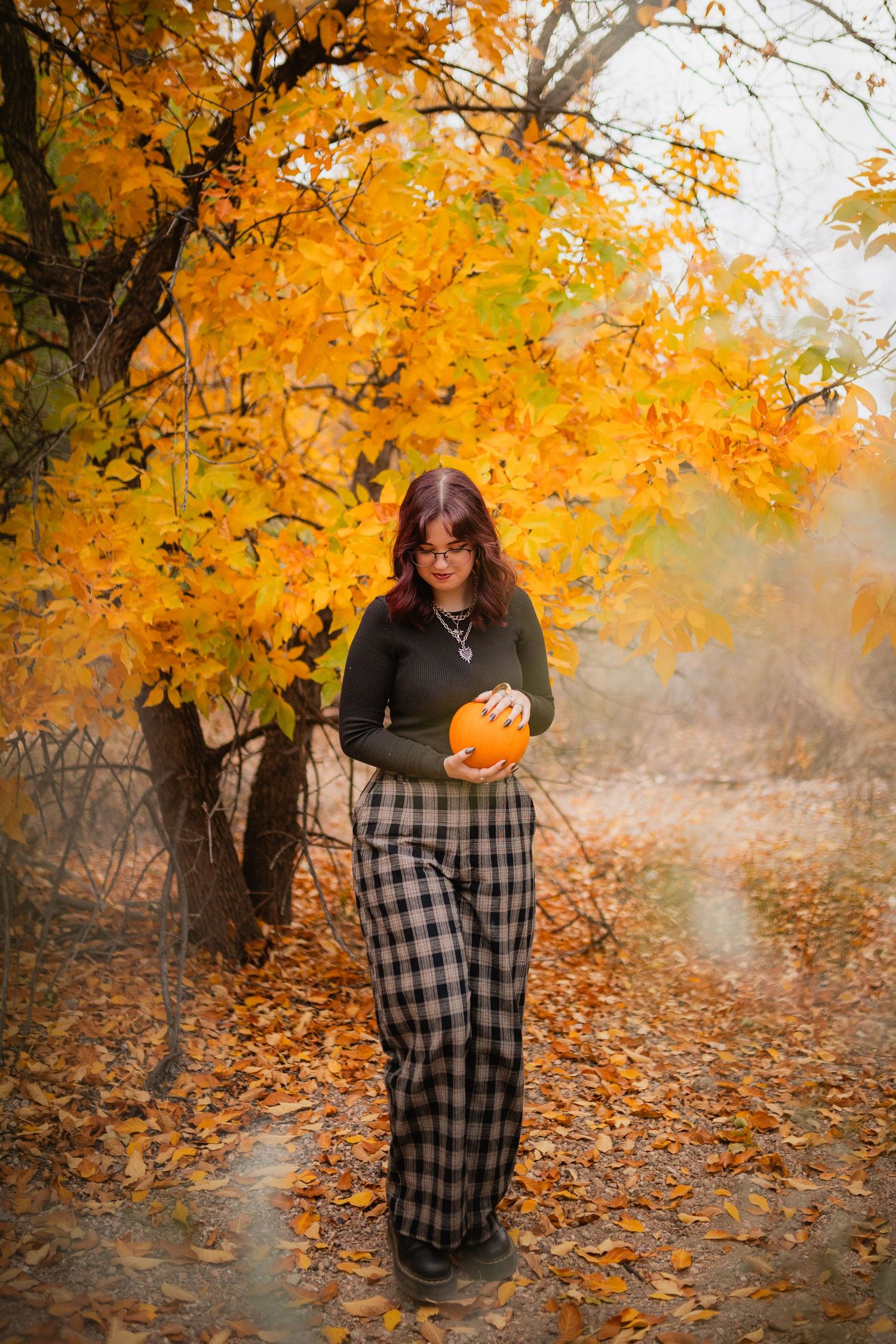 A figure in plaid pants holds a pumpkin under vibrant golden autumn foliage on a misty fall day.