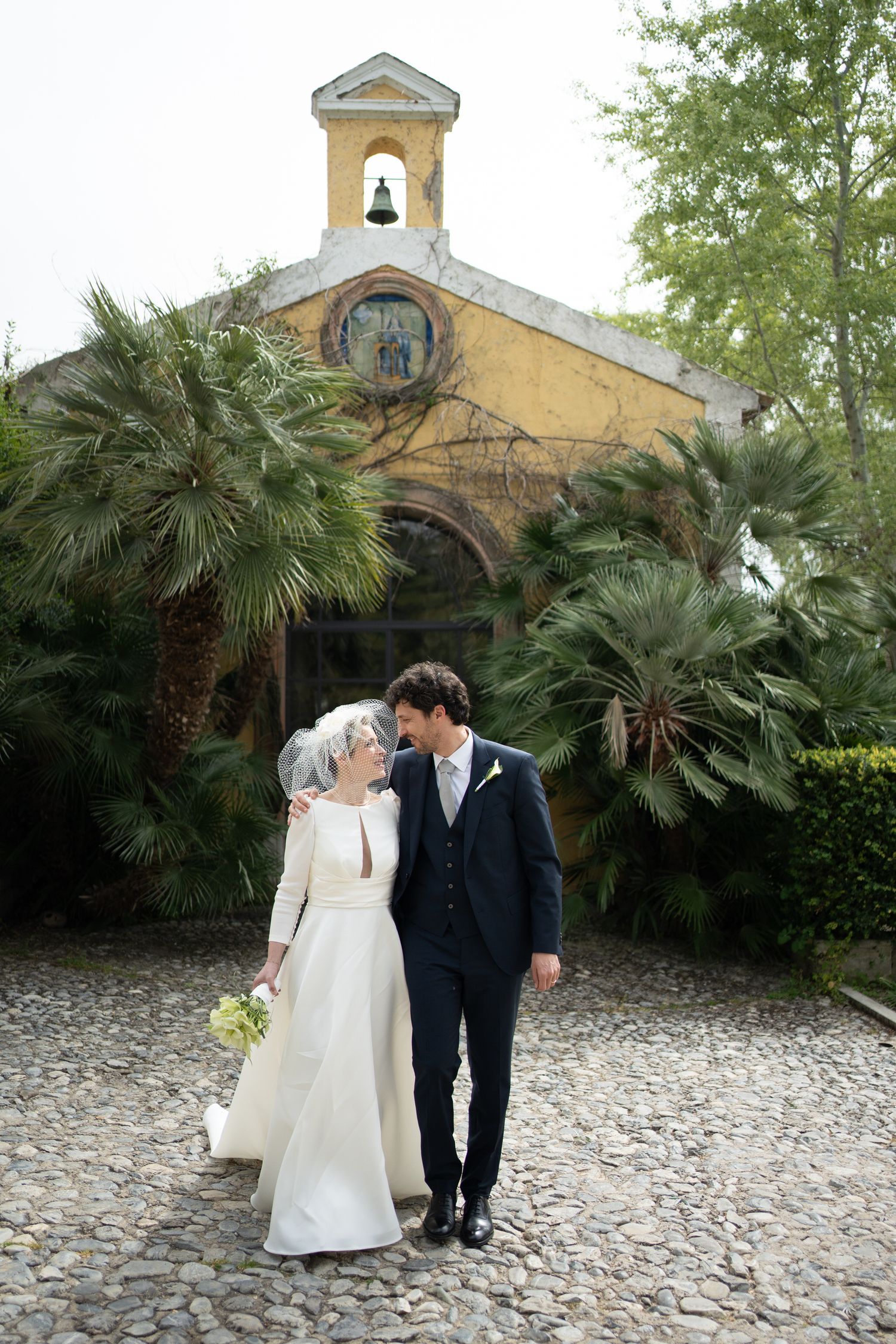 A couple walking along a cobblestone path near a yellow church building with palm trees.
