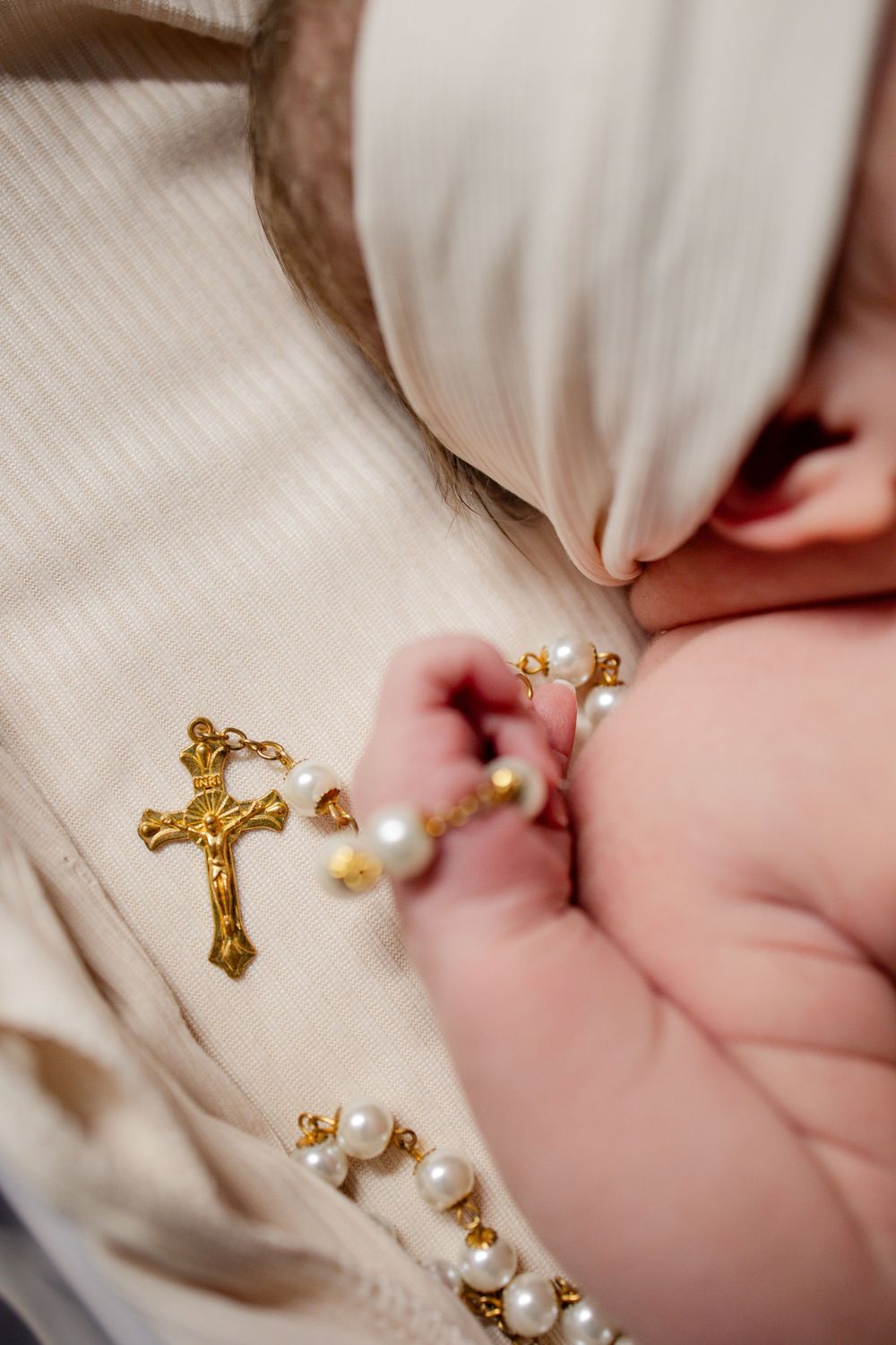 Newborn baby holding a gold crucifix rosary with pearl beads during baptism ceremony.