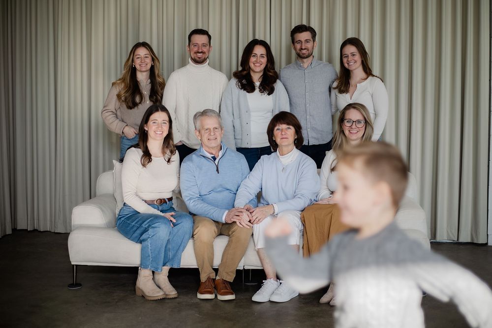 A multigenerational family of ten poses together on a couch in a modern room, smiling warmly, as a toddler runs in the foreground.