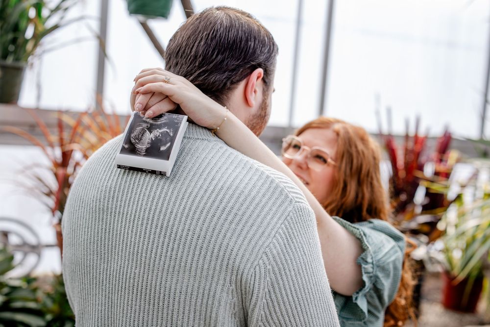 Couple embracing in a greenhouse, woman holding ultrasound photo, pregnancy announcement moment.