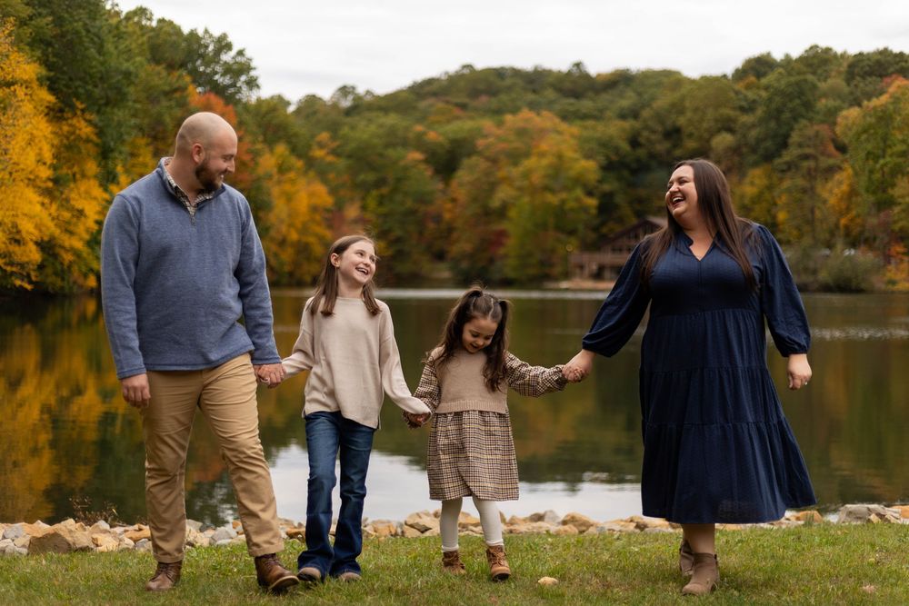 A family walks hand in hand near a lake with vibrant autumn foliage in the background during a fall photo session.