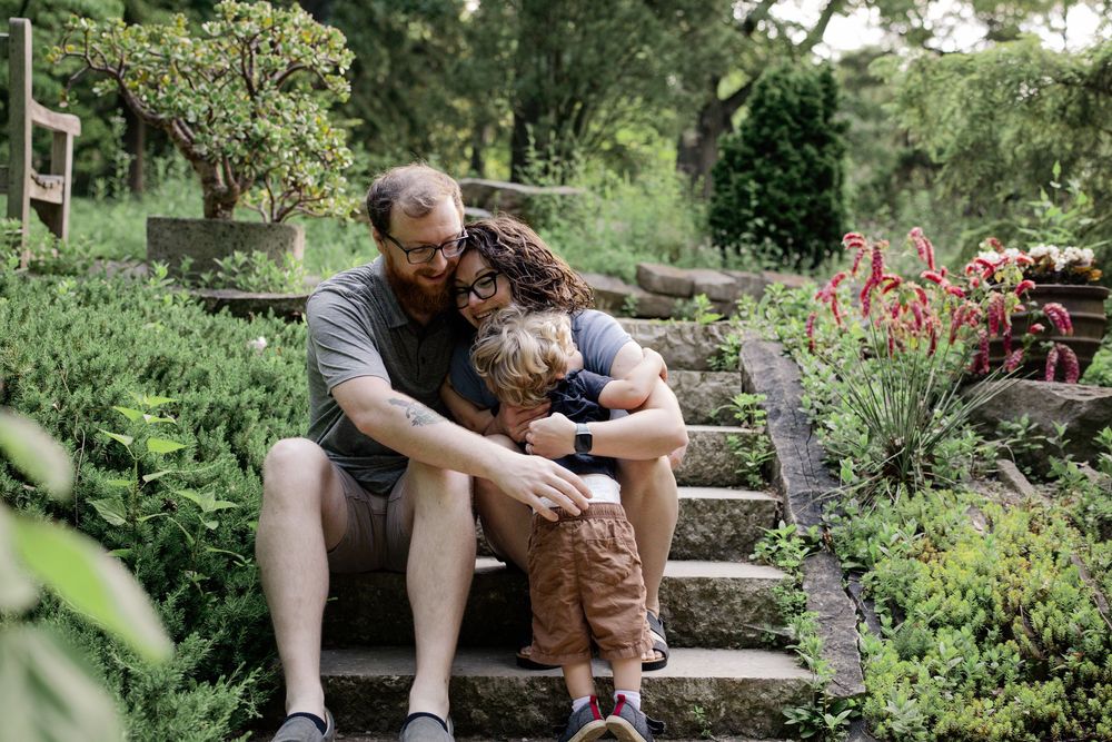 A family of three sits together on stone garden steps surrounded by lush greenery and red flowers.