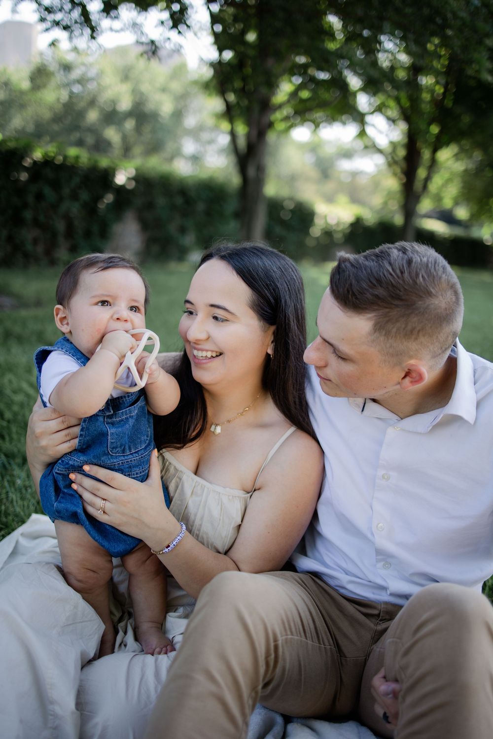 Young family sits together outdoors in a garden setting on a sunny day, with a toddler in denim overalls.