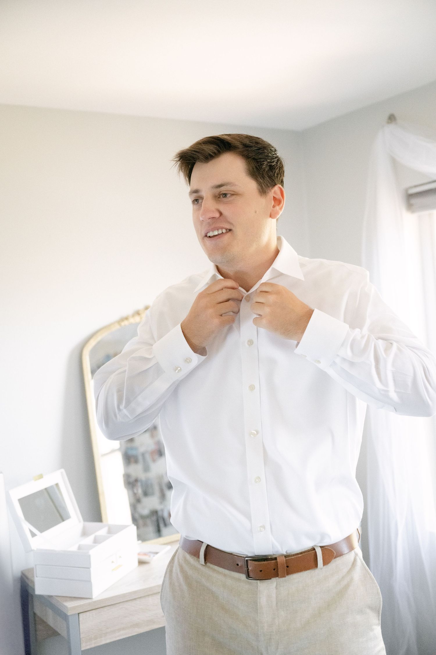 Person in crisp white dress shirt and khaki pants adjusts collar in bright room.