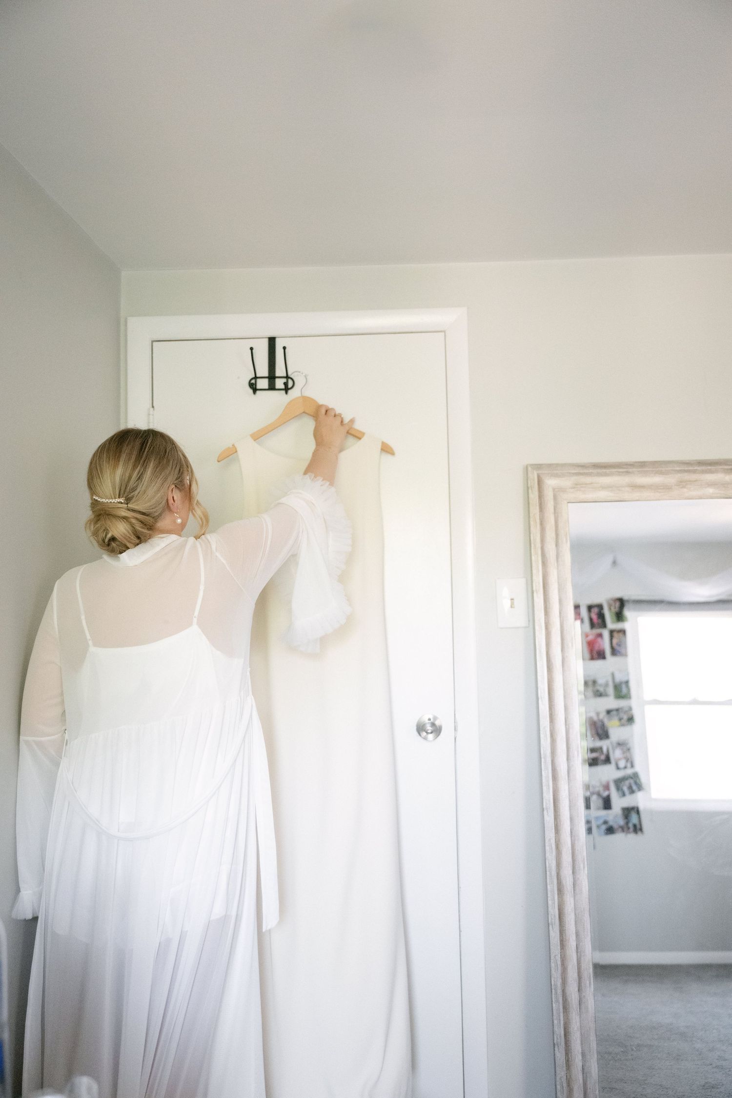 Person in white dress hanging a wedding gown on door hook in bright room.