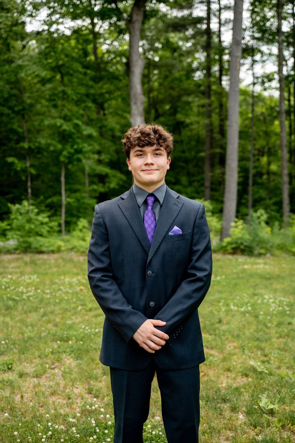 Person in a dark suit with purple tie poses in a green woodland area during formal outdoor photo session.
