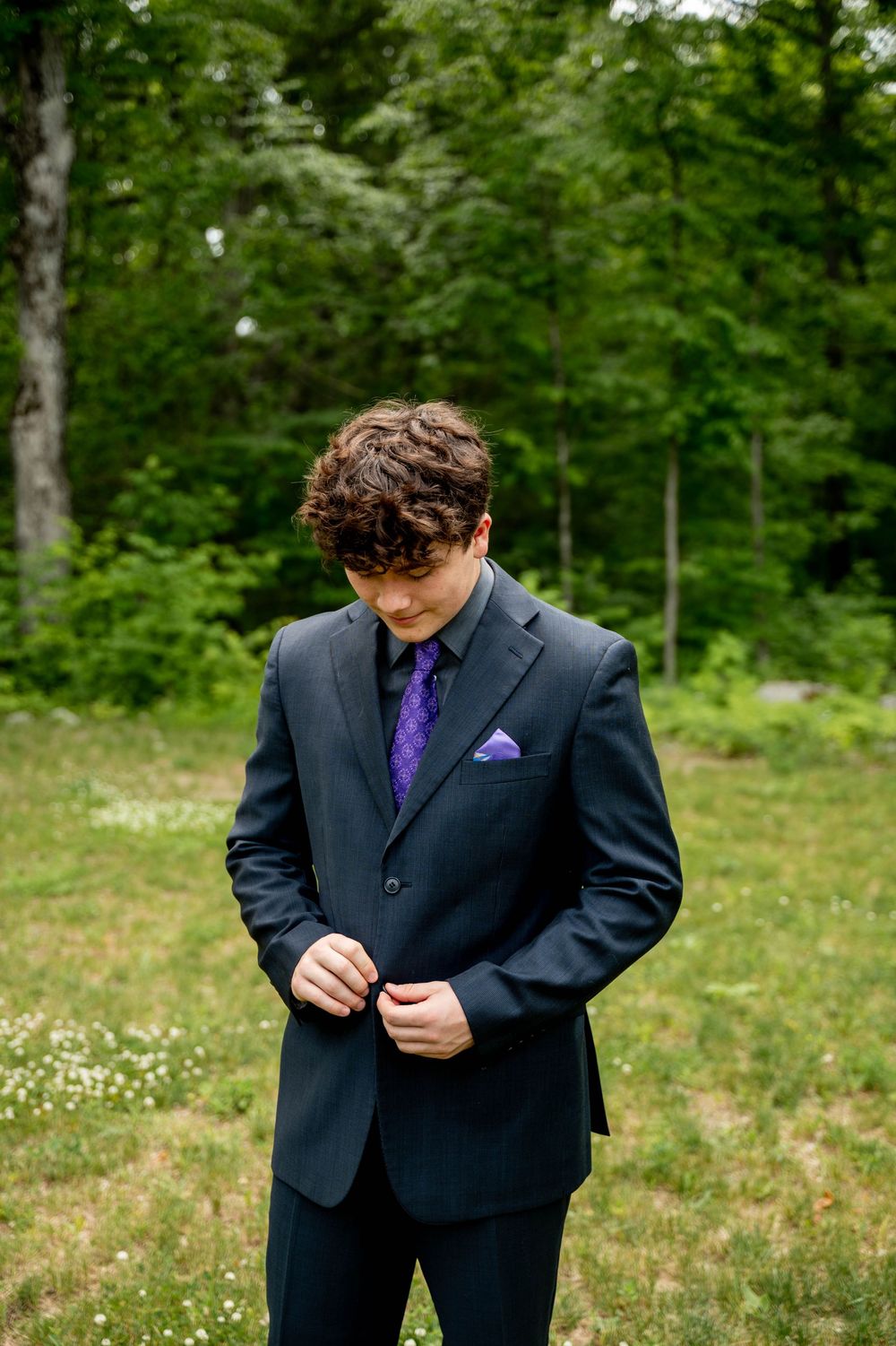 A person in a black suit and purple tie adjusts their clothing while standing in a green outdoor setting.