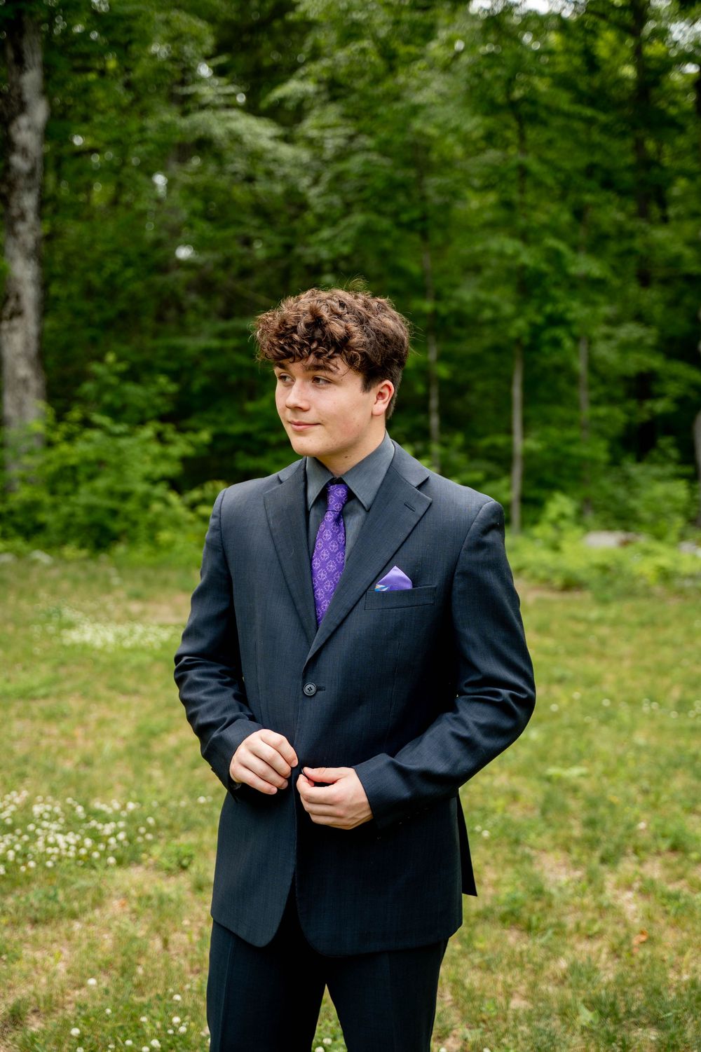 Sharp navy suit with purple tie and pocket square ensemble photographed outdoors against lush green forest backdrop.