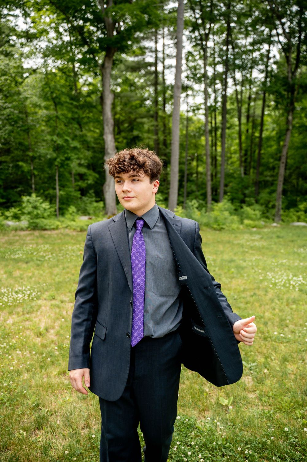A person wearing a dark suit and purple tie poses expressively in a sunny meadow with trees in the background.