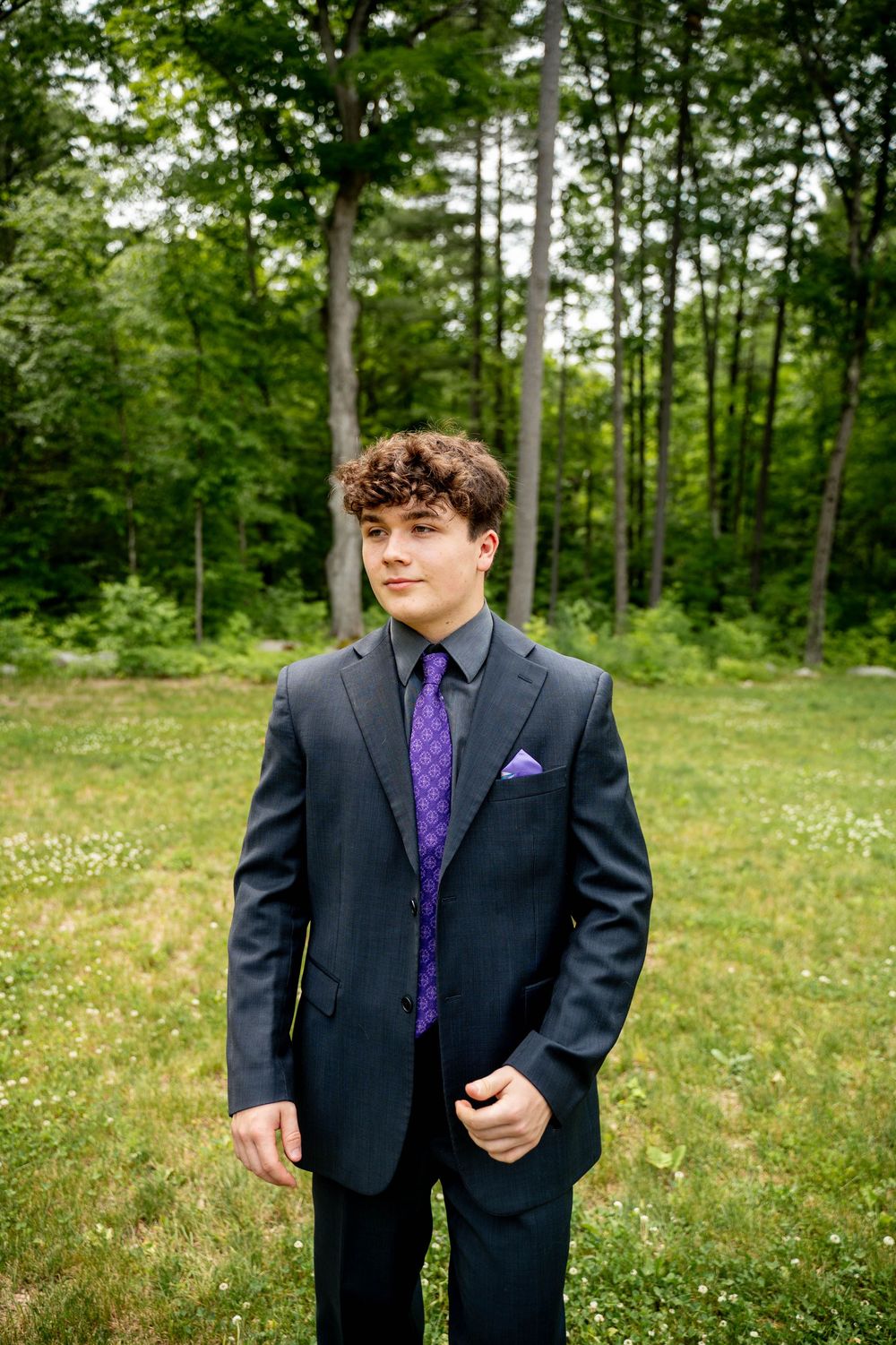 Formal portrait of someone wearing a charcoal suit with purple tie and pocket square in a lush green forest setting.