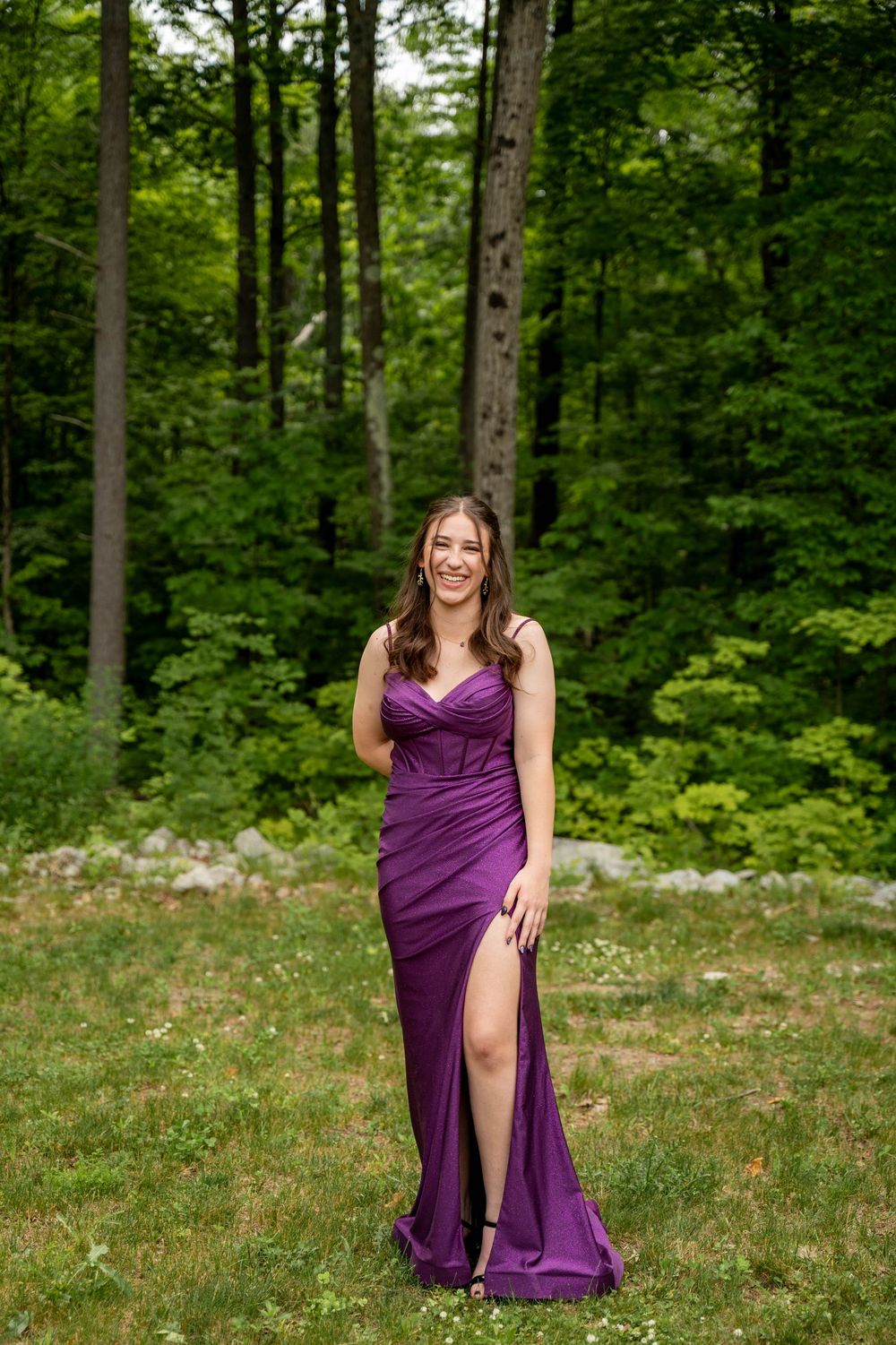 An elegant purple formal gown with a side slit stands out against the lush green forest backdrop on a summer day.