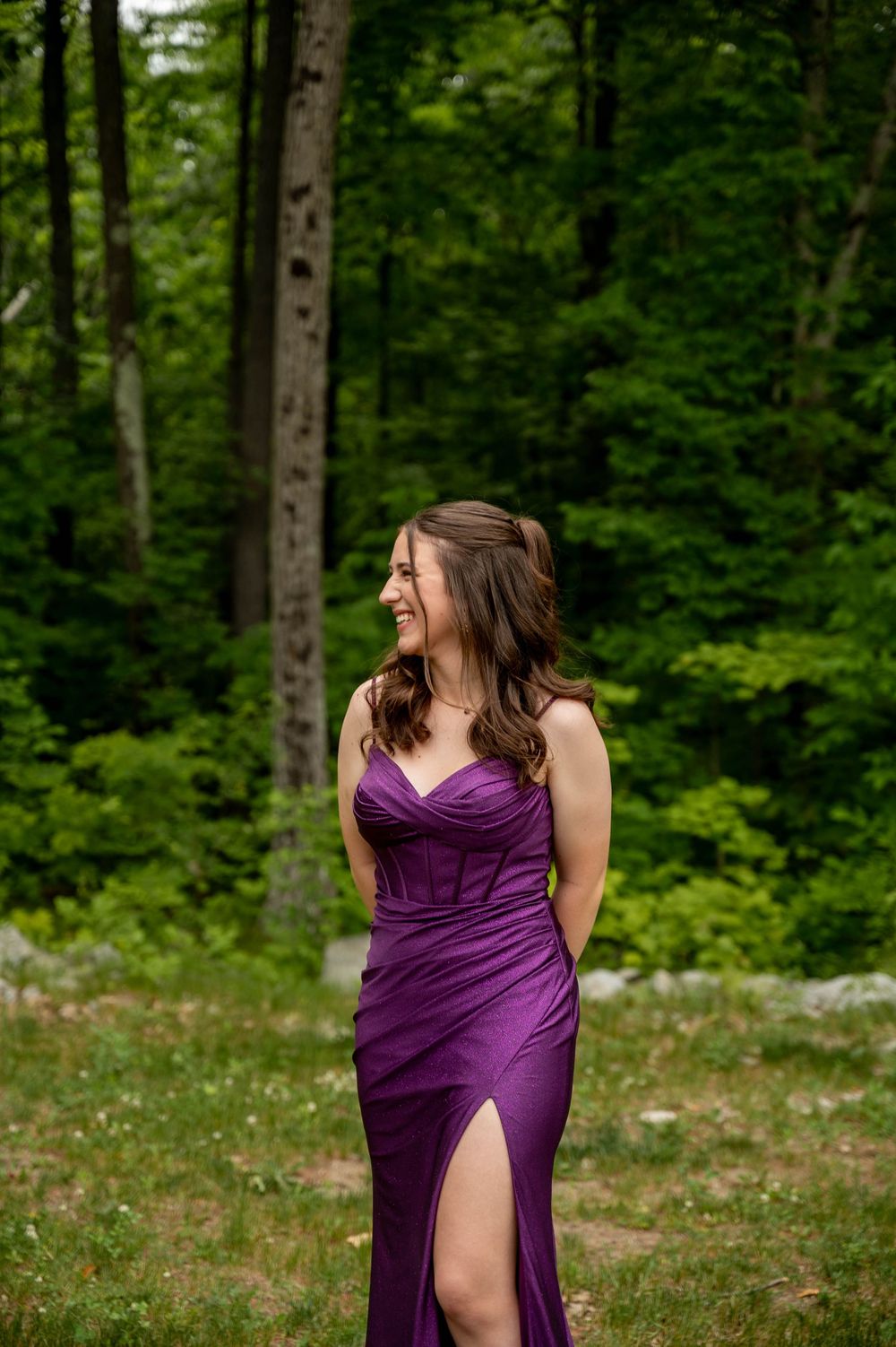A long purple formal dress with a slit stands out against a lush green forest background.