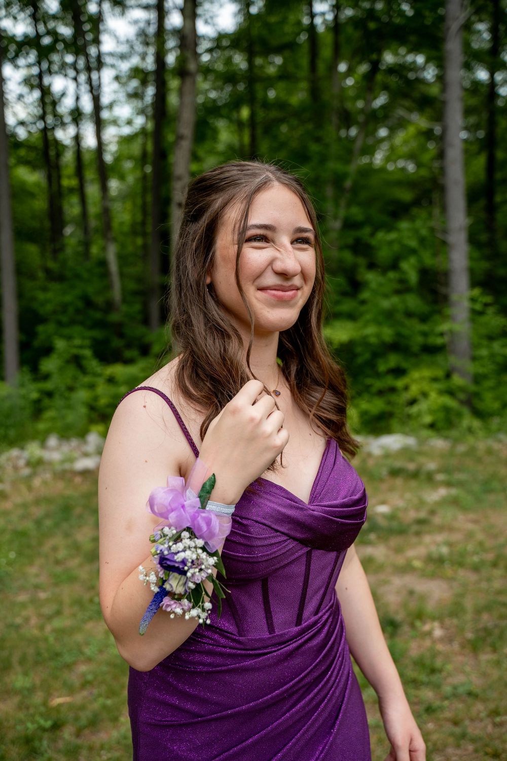 A person in a purple bridesmaid dress with a floral wrist corsage smiles in a wooded outdoor setting.