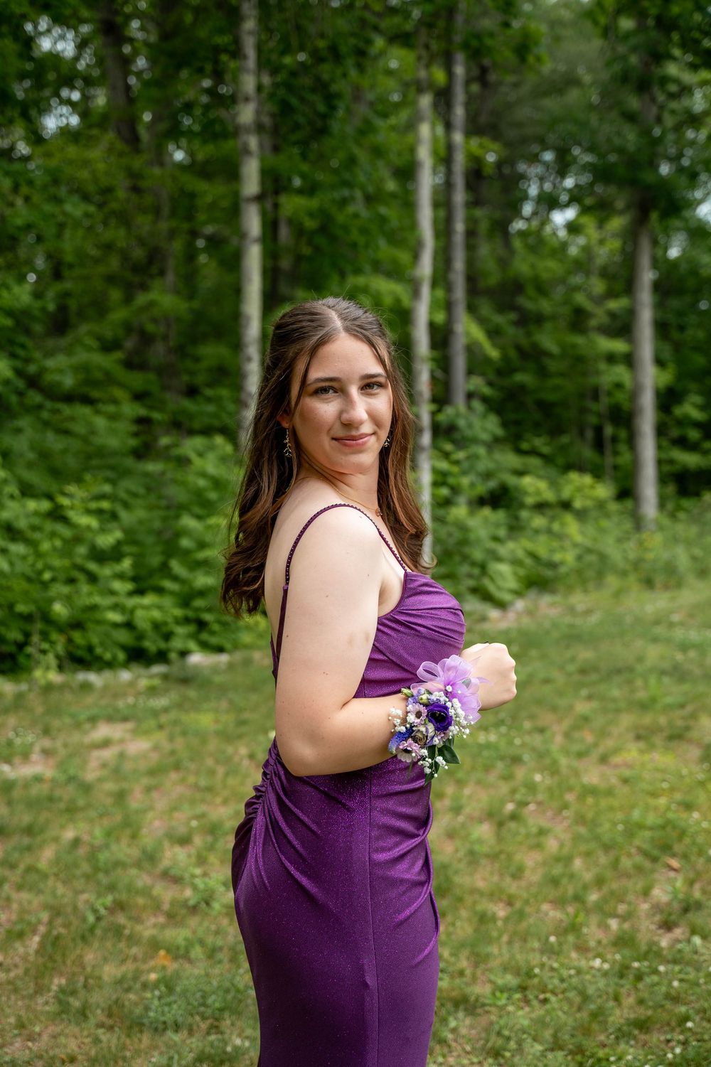 A person wearing a purple formal dress stands in a wooded outdoor setting holding a floral corsage.