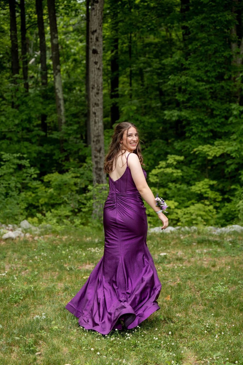 A person in a flowing purple formal gown twirls in a lush green forest setting with wildflowers in the grass.
