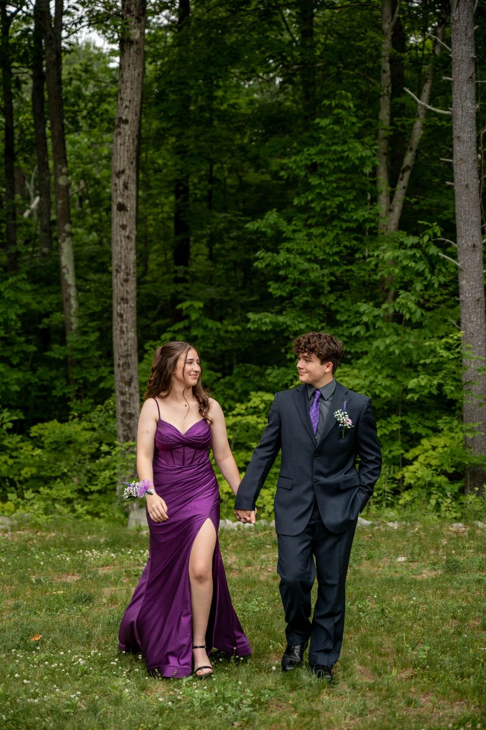 Couple in purple dress and navy suit share romantic moments in a lush forest setting during formal event.