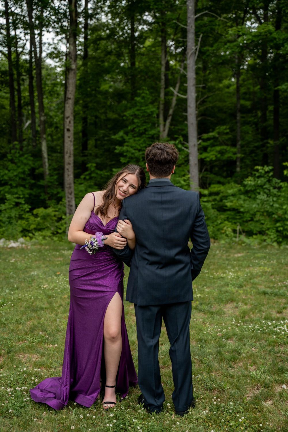 A couple in formal attire stands on grass with lush green forest trees in the background.