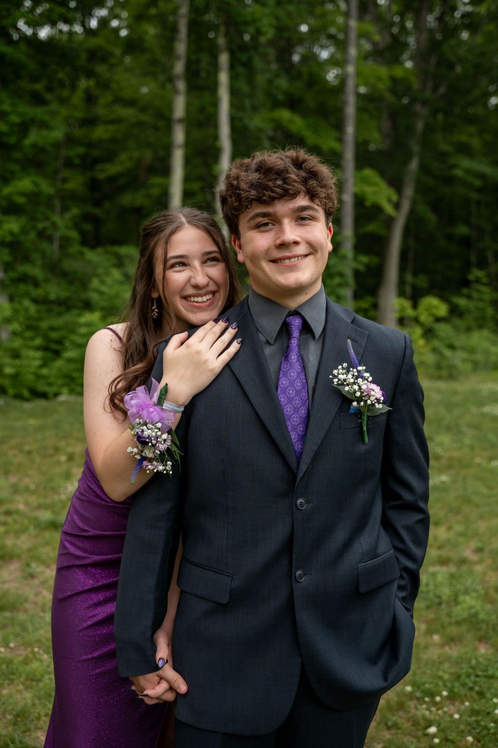 A formal couple dressed in purple and black attire poses together outdoors in a natural woodland setting.