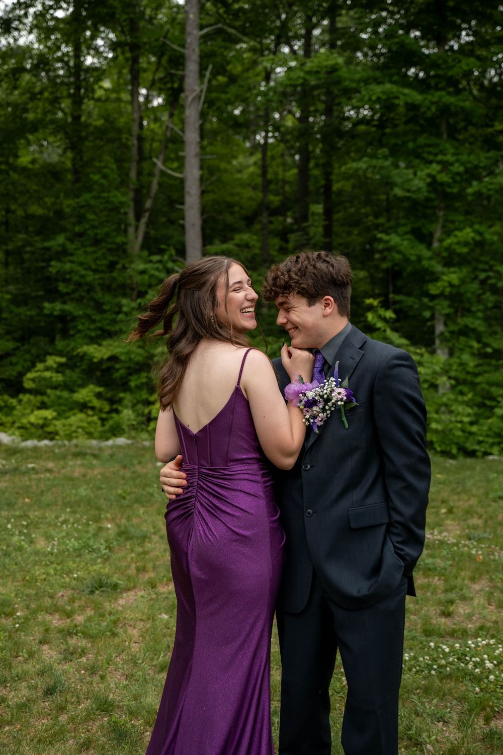 A couple shares a joyful moment outdoors in formalwear, with her in a purple dress and him in a dark suit.