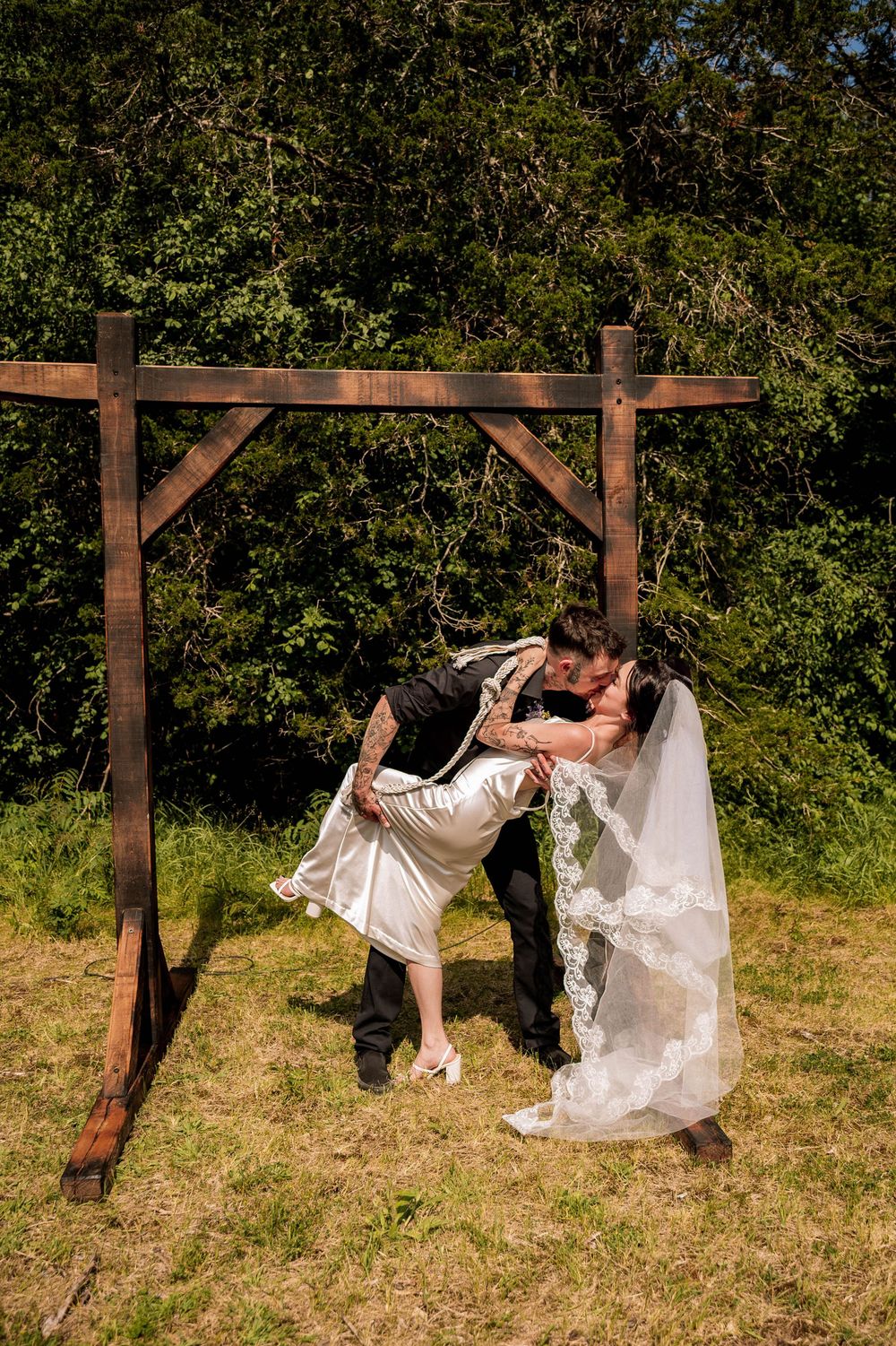 A couple shares a playful dip pose under a wooden arch at an outdoor wedding.