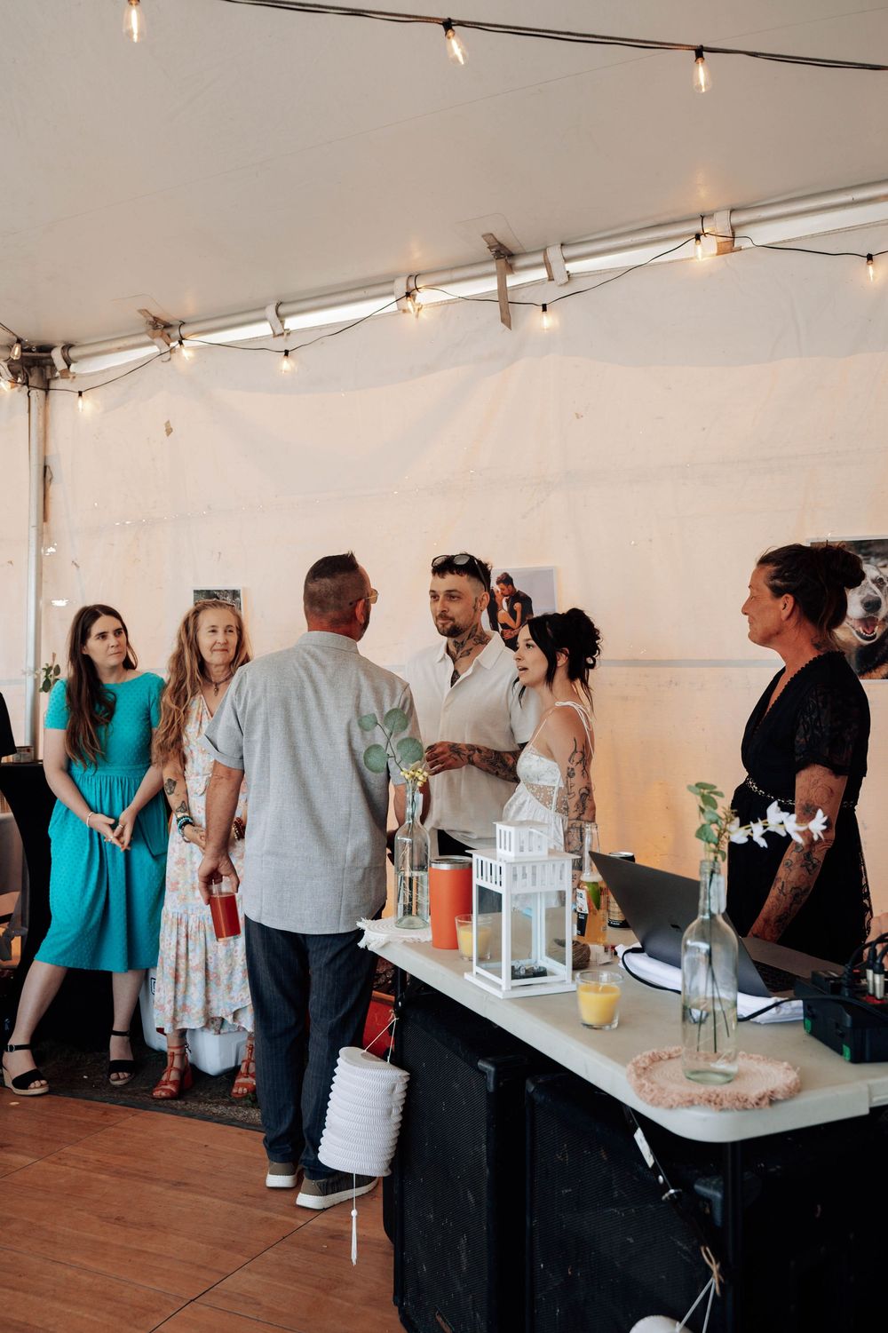 Guests enjoying refreshments at an outdoor wedding reception under a white tent with string lights.