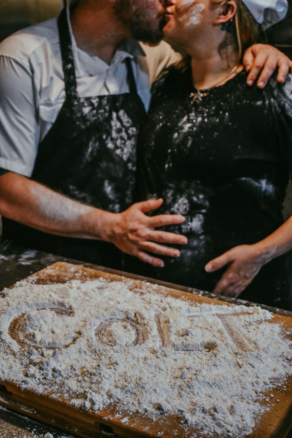 Creative maternity photo with baby name in flour and expectant parents dressed as chefs in background.