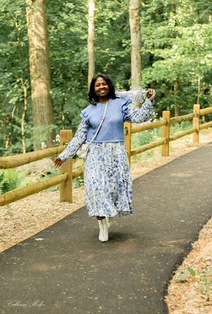 Woman in flowing blue floral dress poses elegantly along a tree-lined path in a serene wooded setting.