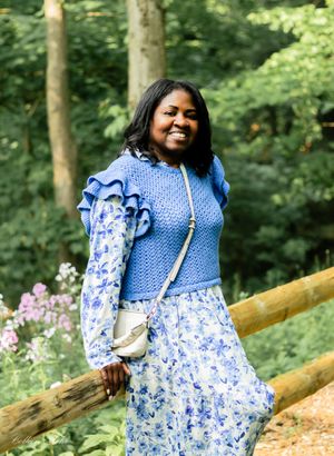 A person wearing a blue and white floral dress stands on a wooden bridge in a lush forest setting.