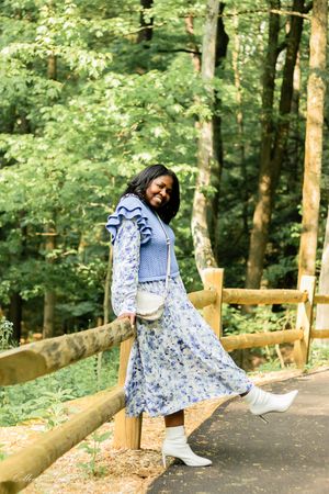 A person in a blue floral dress and white boots poses playfully along a wooden railing on a forest path.