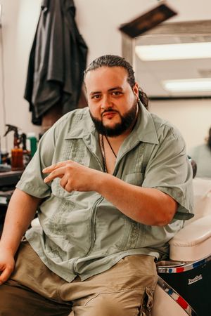 A person in a casual olive button-up shirt and khaki pants sits indoors in a relaxed pose.