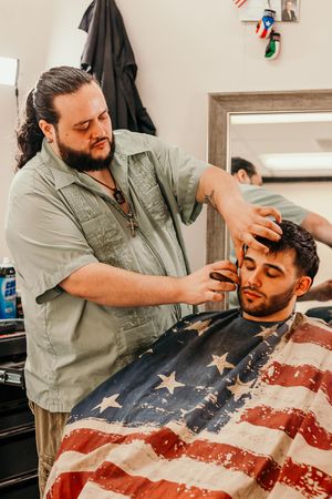 A barber performs a haircut on a client draped in an American flag-styled cape in a modern barbershop setting.