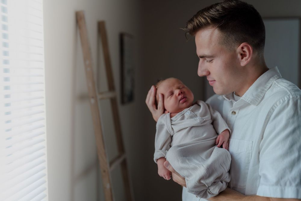 A parent dressed in a white shirt tenderly holds and gazes at a newborn baby swaddled in white at an in-home newborn session in Ohio