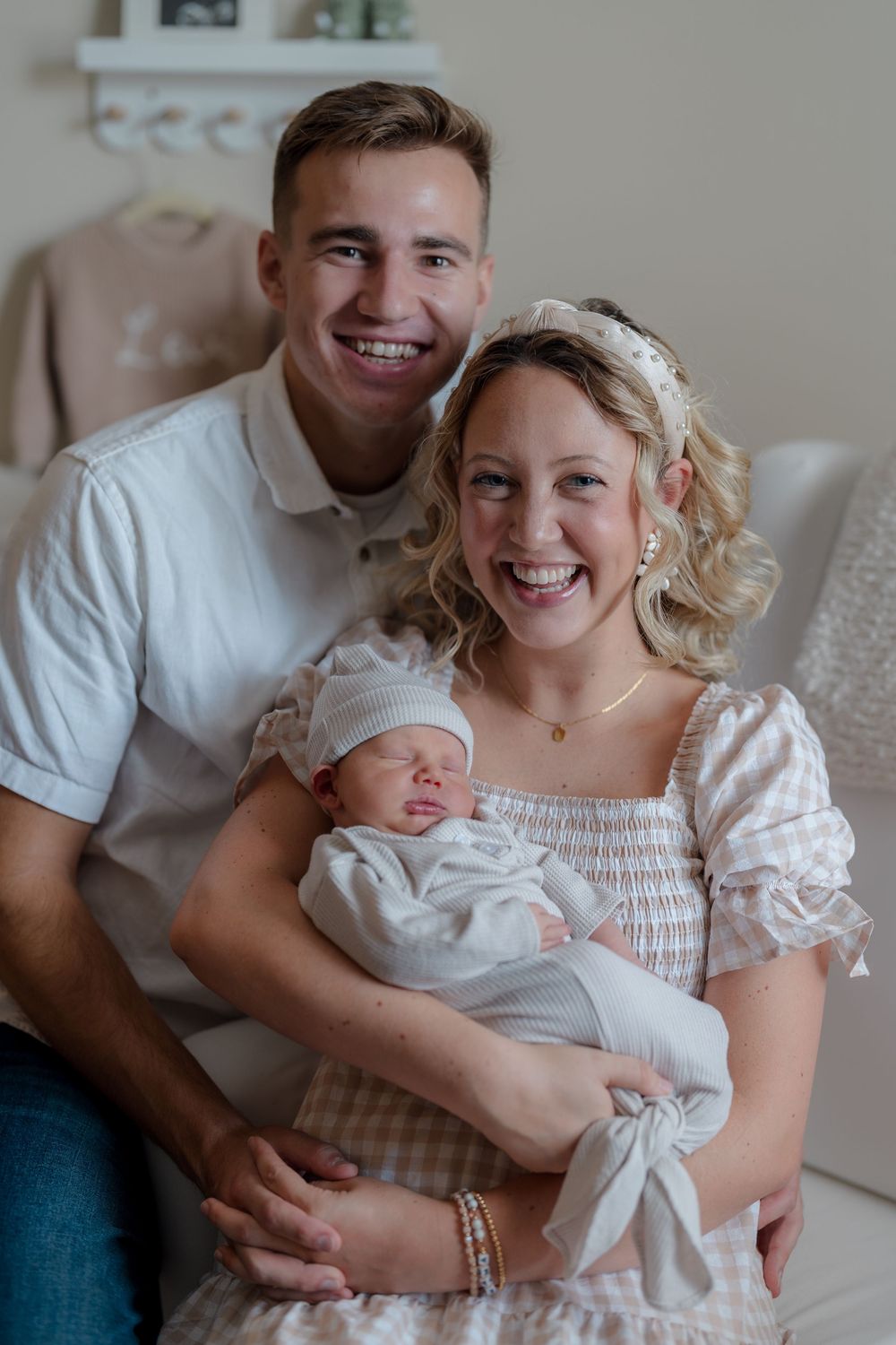 A happy family portrait shows parents in white outfits sitting on a chair with their sleeping newborn at Cuyahoga Falls, Ohio in-home photo