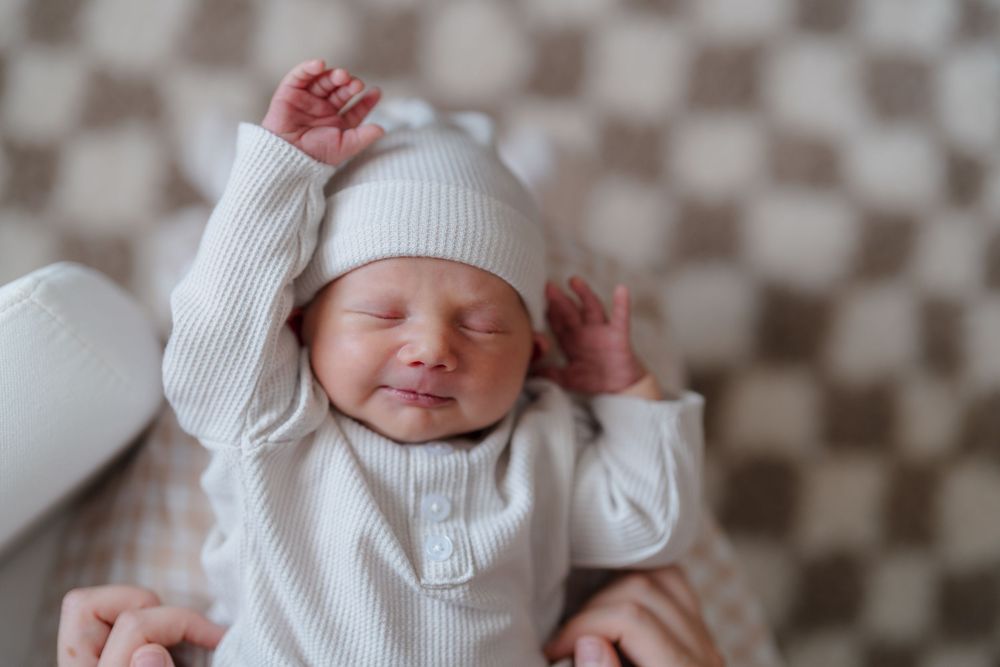 A newborn baby smiles while wearing a white knit hat and swaddle blanket on a checkered background at in-home session in Cuyahoga Falls, OH