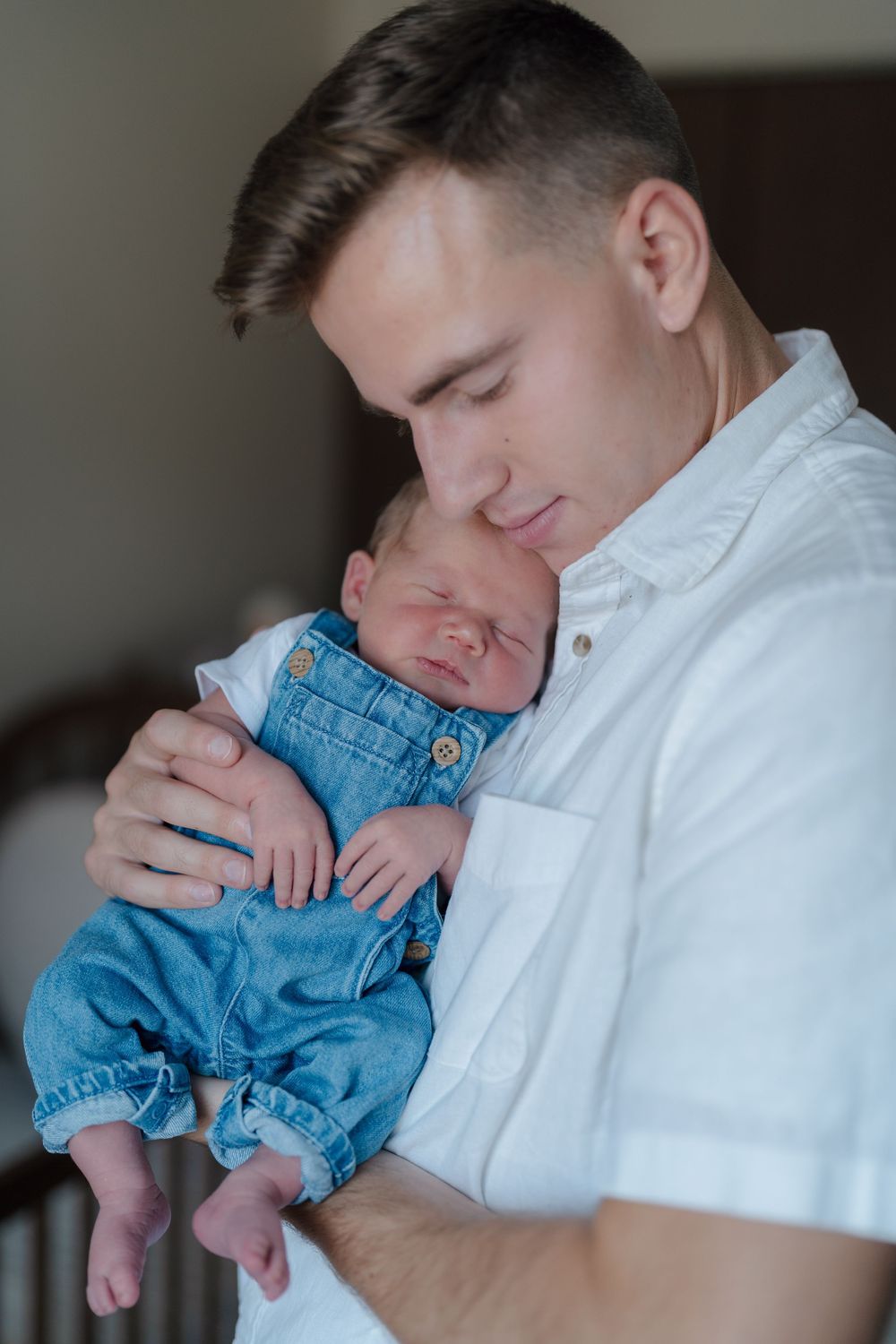 A tender moment shows someone in a white shirt holding a sleeping baby wearing blue denim overalls at in-home newborn session near Stow, OH