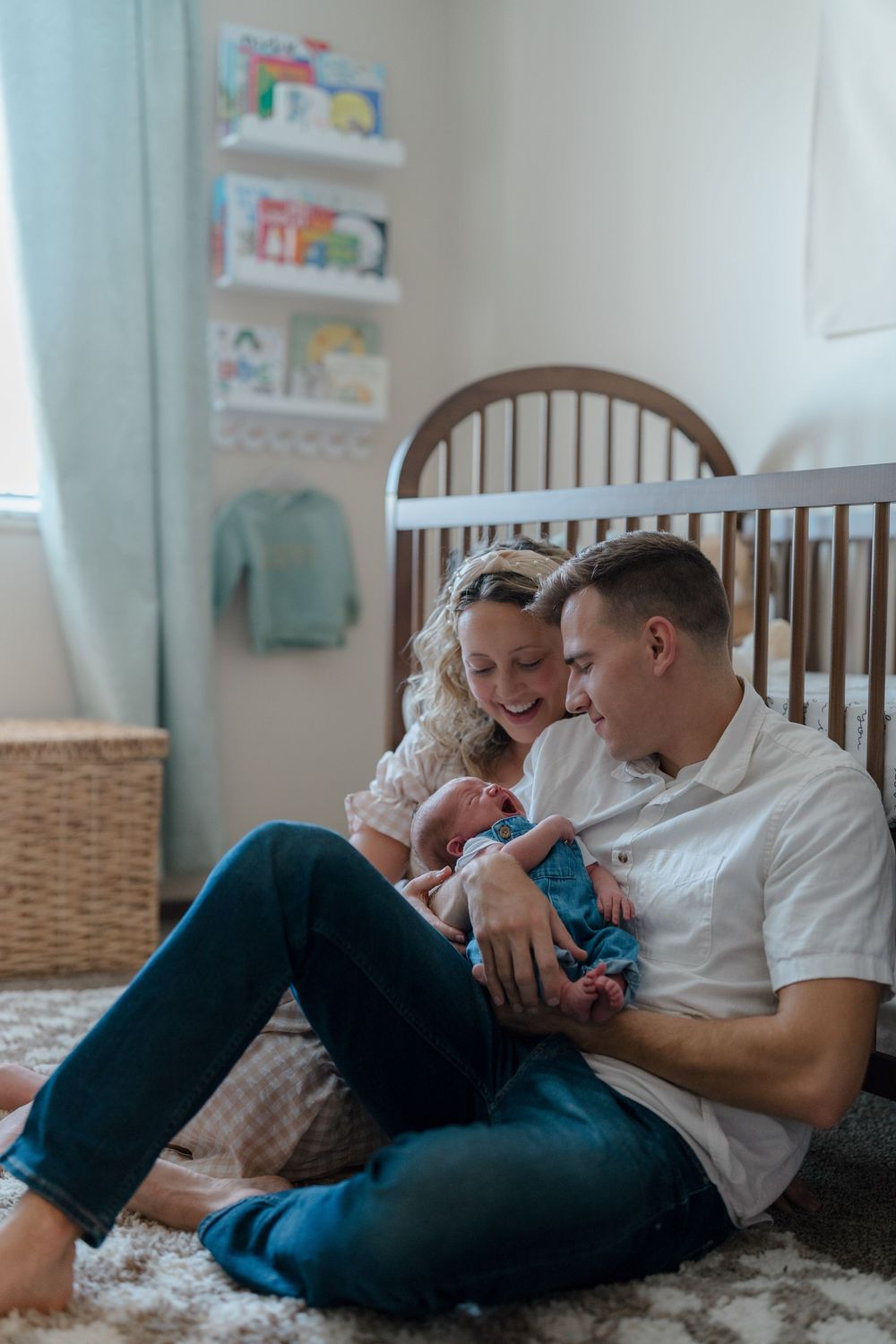 A couple sits together on the floor of a cozy nursery with their newborn baby next to a wooden crib at in-home newborn photo session in Ohio