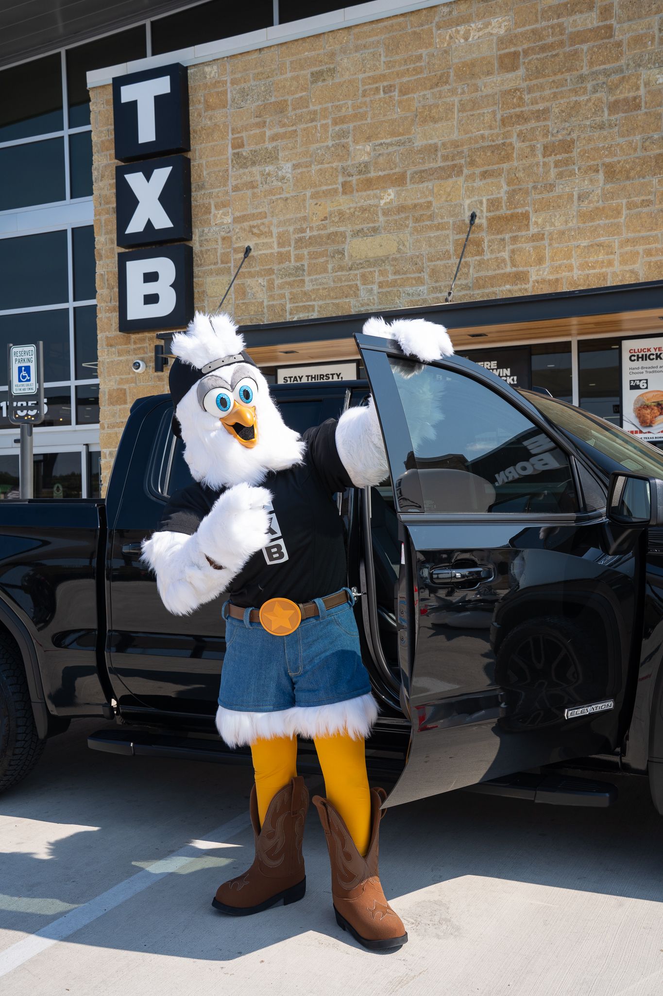 A person in an eagle mascot costume poses in front of a brick building and parked cars.