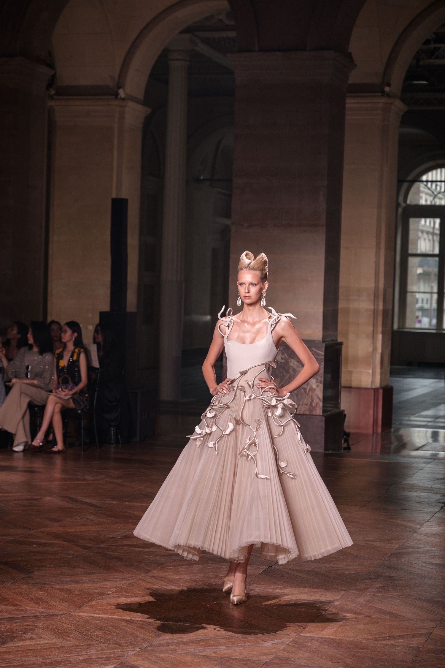 A model twirls in a flowing beige princess-style dress during a photoshoot in an elegant stone archway corridor.