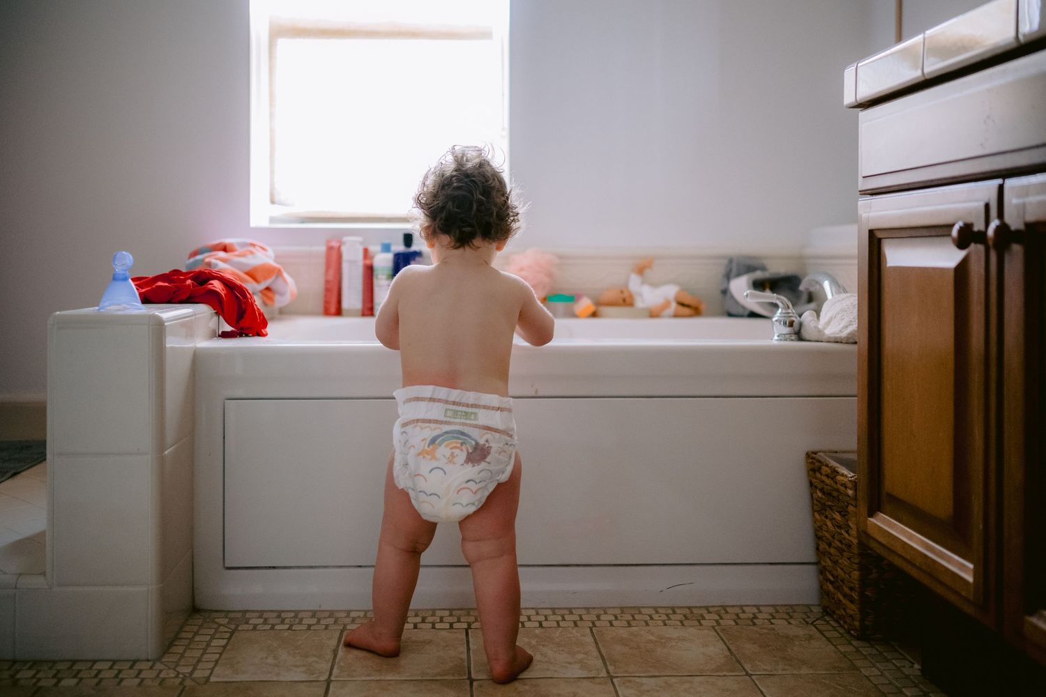 A toddler in a white diaper standing along the edge of a white bathtub.