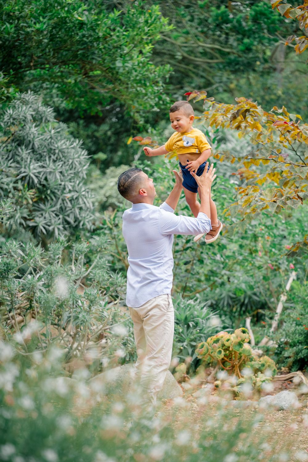 A parent lifting a small child wearing a yellow shirt in a lush green garden in Huntington Beach.