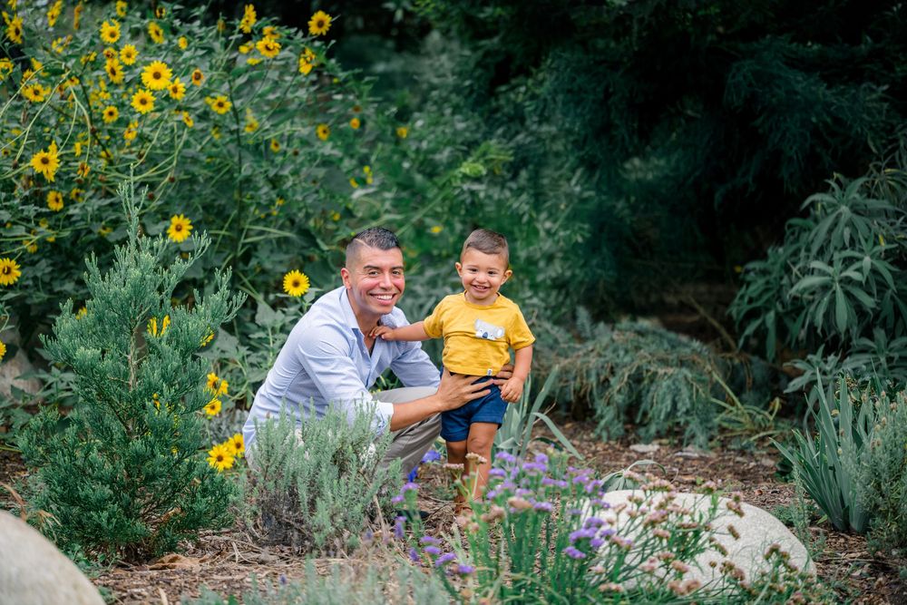 Father and son smiling in bright yellow sunflowers and greenery in a garden in Huntington Beach, California.