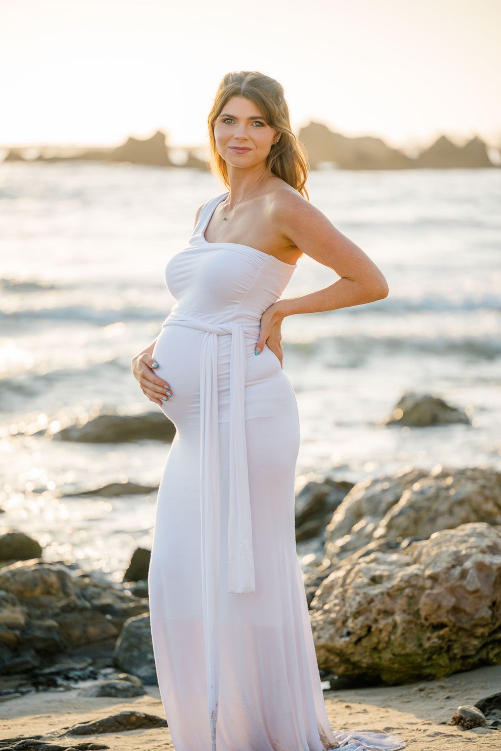 Pregnant mother in a white flowy dress holds her belly during golden hour maternity photo session at Newport Beach.