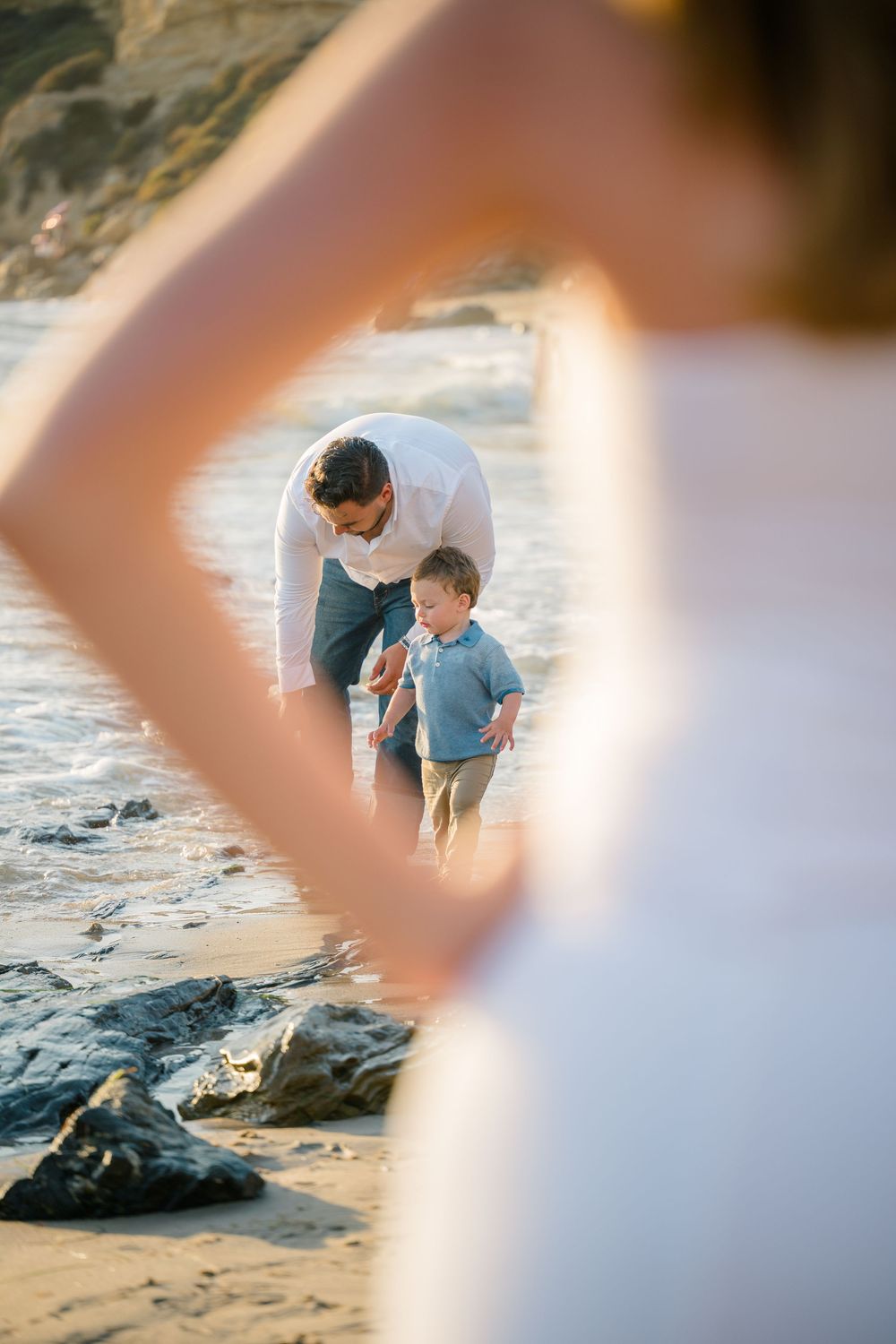 Creative beach family photography framed through hands showing father and son walking together along shore in Newport Beach at sunset.