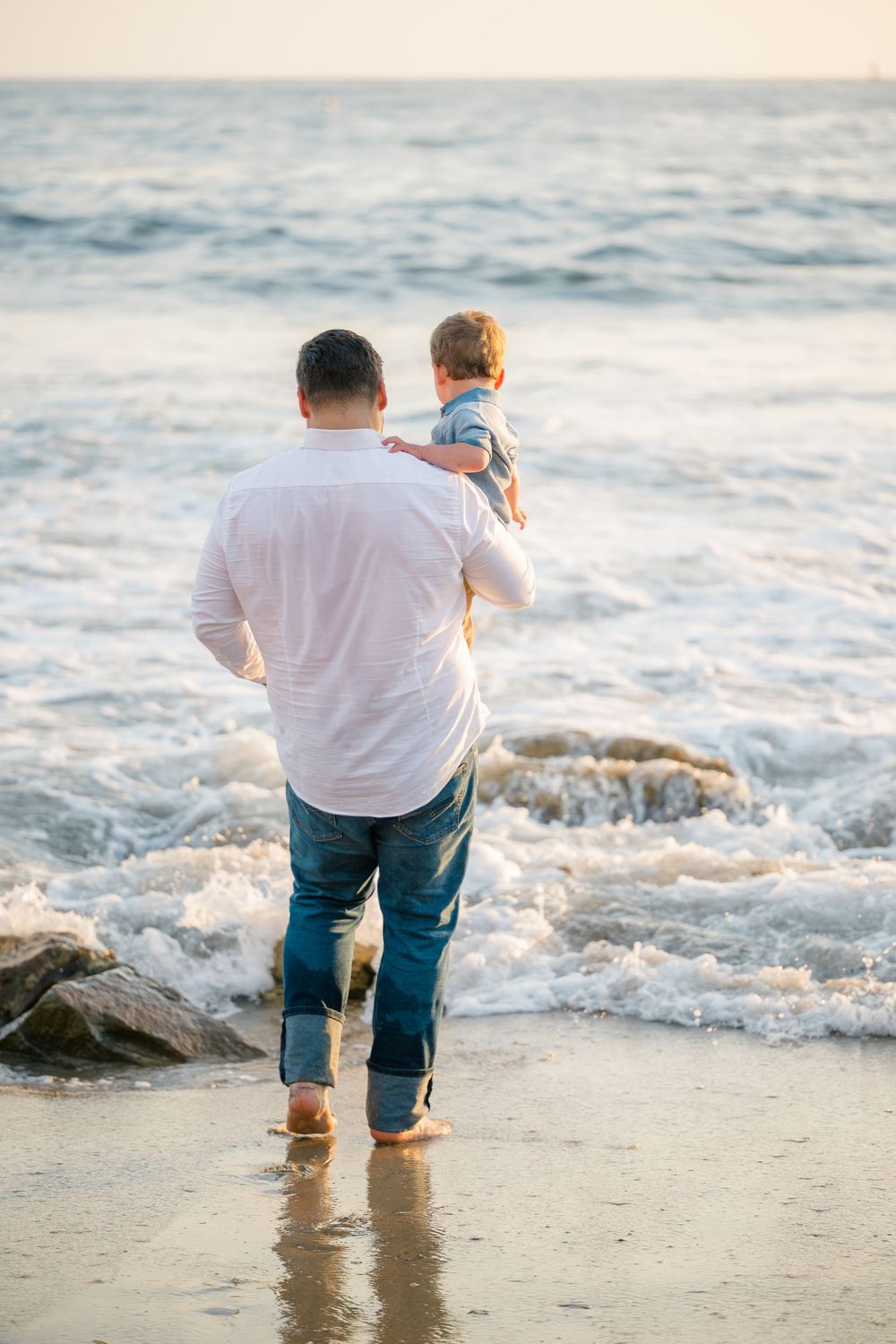 Father carries with his toddler son at the water’s edge during a Newport Beach family lifestyle photo session in Orange County.