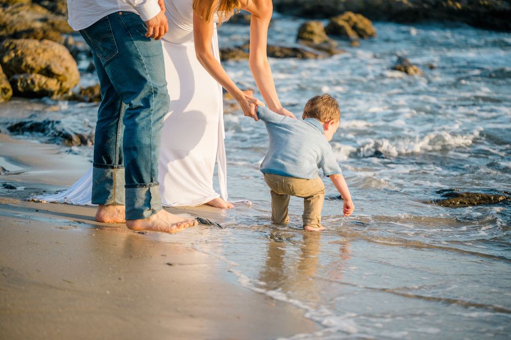 A small child plays at the water's edge on Newport Beach with parents for family photography session at sunset in Orange County.
