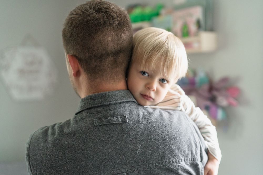 A small child peeks over the shoulder of an adult wearing a gray shirt in a softly lit room at the in home newborn photo session in Ohio