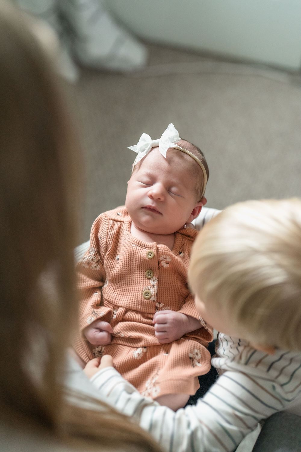 Sleeping baby wearing peach-colored outfit with white bow headband on couch with brother at in home family session in Cuyahoga Falls, Ohio